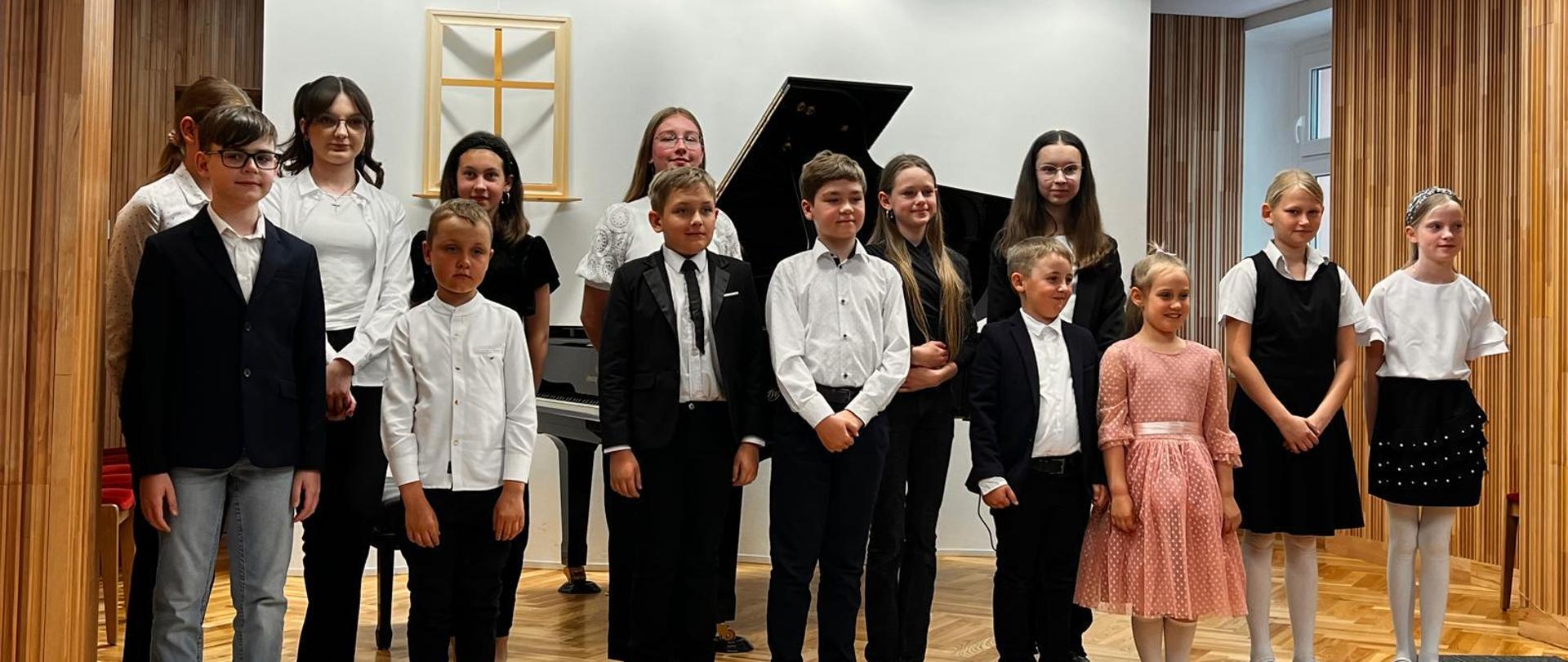 Zdjęcie przedstawia wykonawców II Międzyszkolnej Audycji Fortepianowej stojący grupowo na scenie, za nimi fortepian