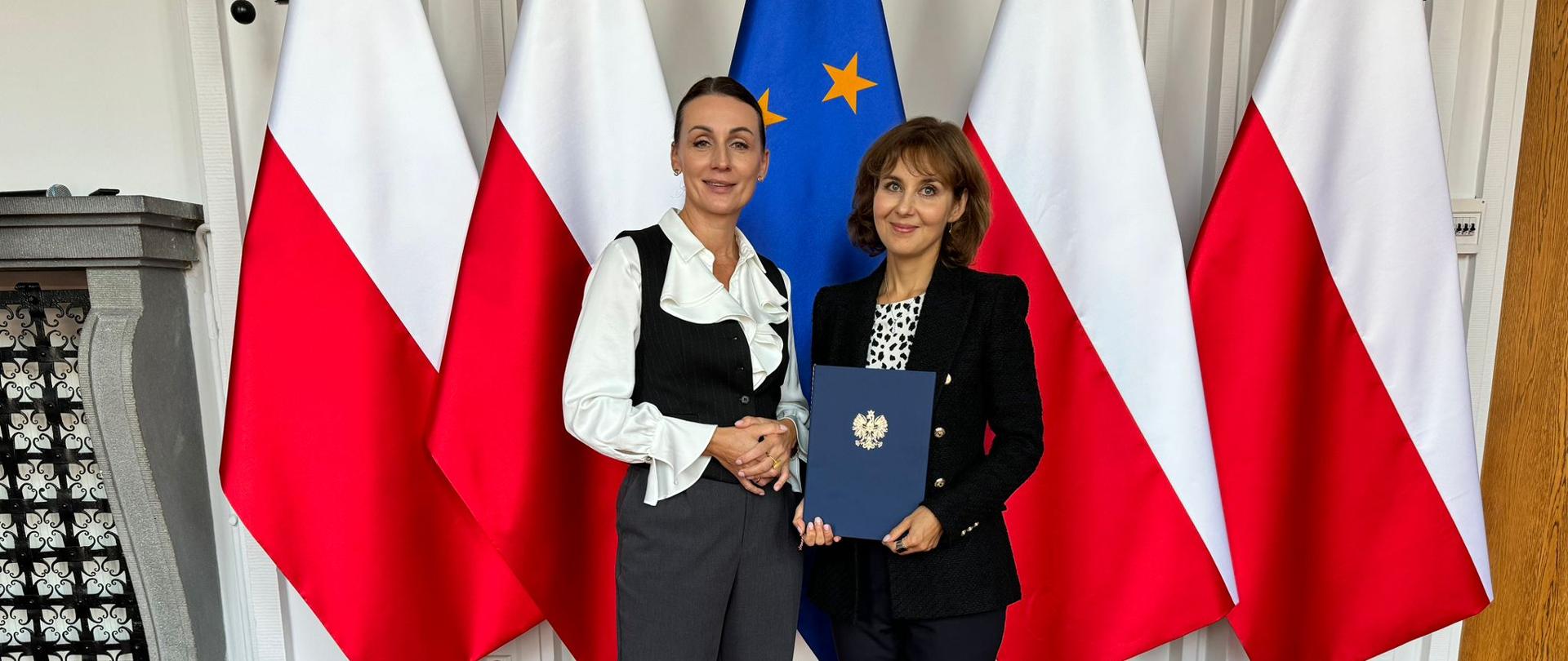 Dwie kobiety stojące na tle czterech polskich flag i jednej flagi Unii Europejskiej. Kobieta po prawej stronie trzyma w dłoniach granatową teczkę. Obie są elegancko ubrane. 