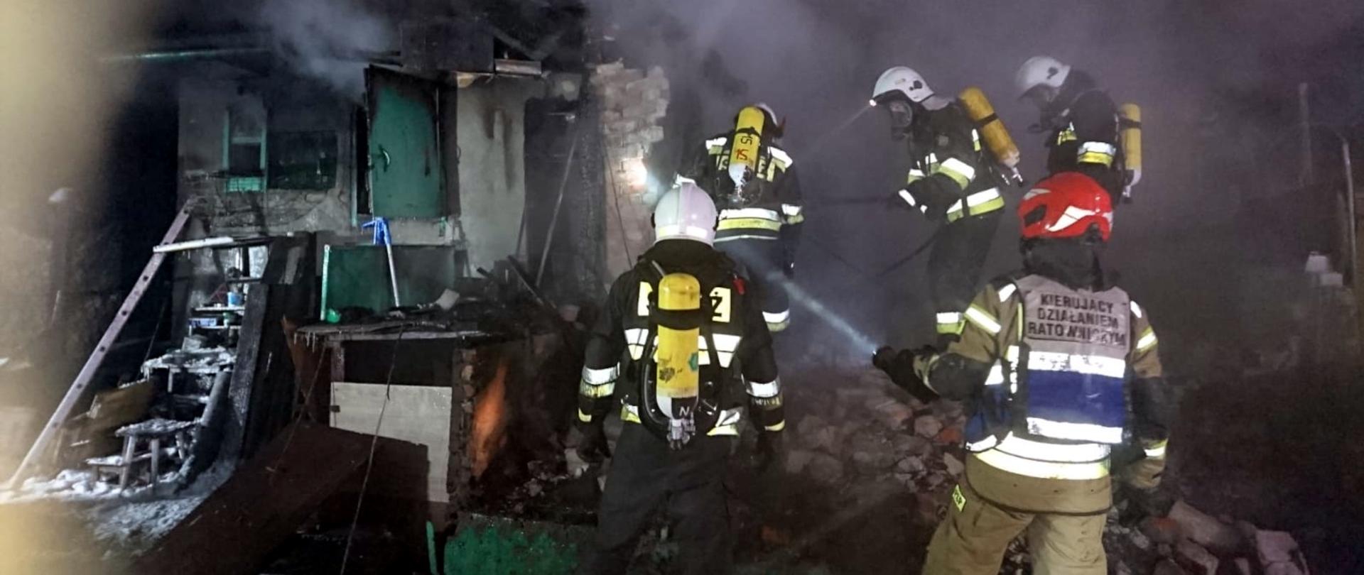 Zdjęcie przedstawia pięciu strażaków w ubraniach specjalnych, hełmach oraz aparatach ochrony układu oddechowego prowadzących działania gaśnicze na zgliszczach altany.