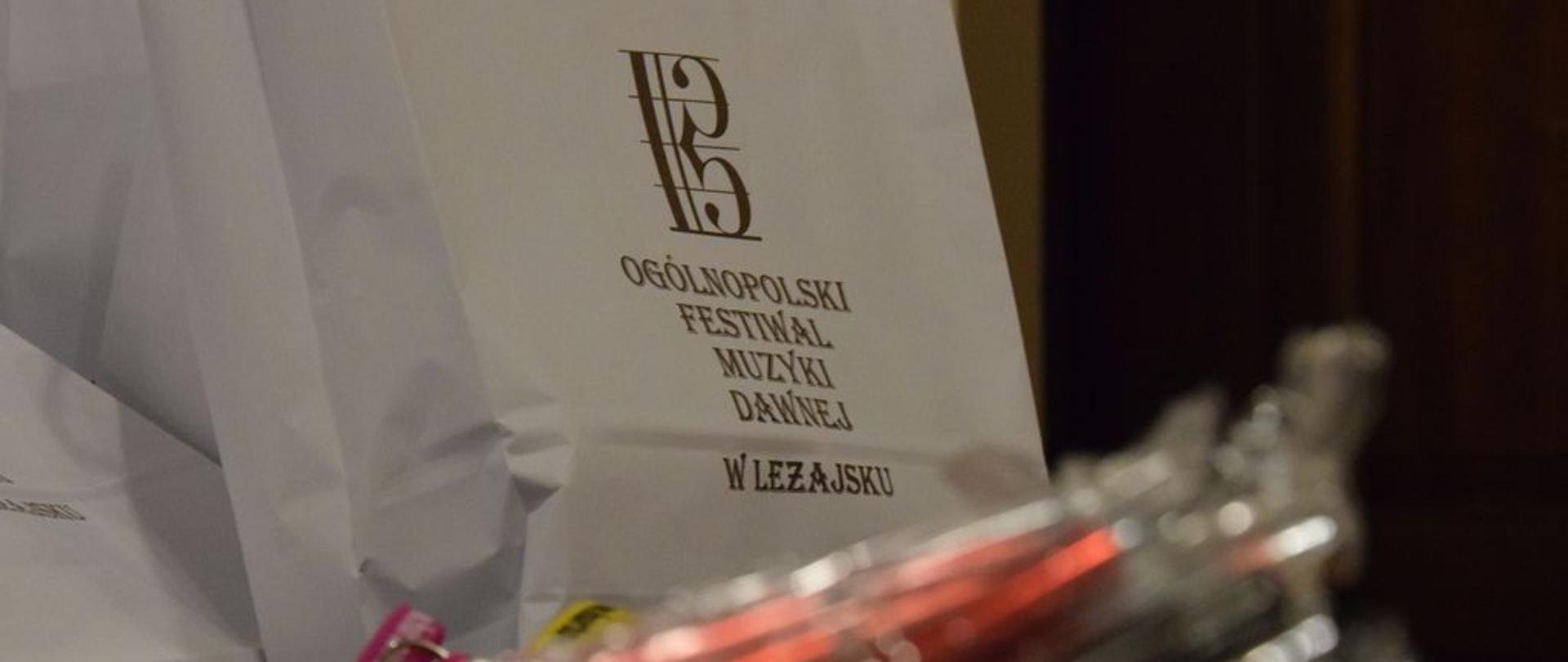 Nagrody dla uczestników Ogólnopolskiego Festiwalu Muzyki Dawnej. Zdjęcie utrzymane w lekkich jasnych barwach. Na zdjęciu na pierwszym planie rozmazany zarys długopisów w kolorze czerwonym, dale torby białę z nagrodami - na torbach brązowy napis z nazwą festiwalu.