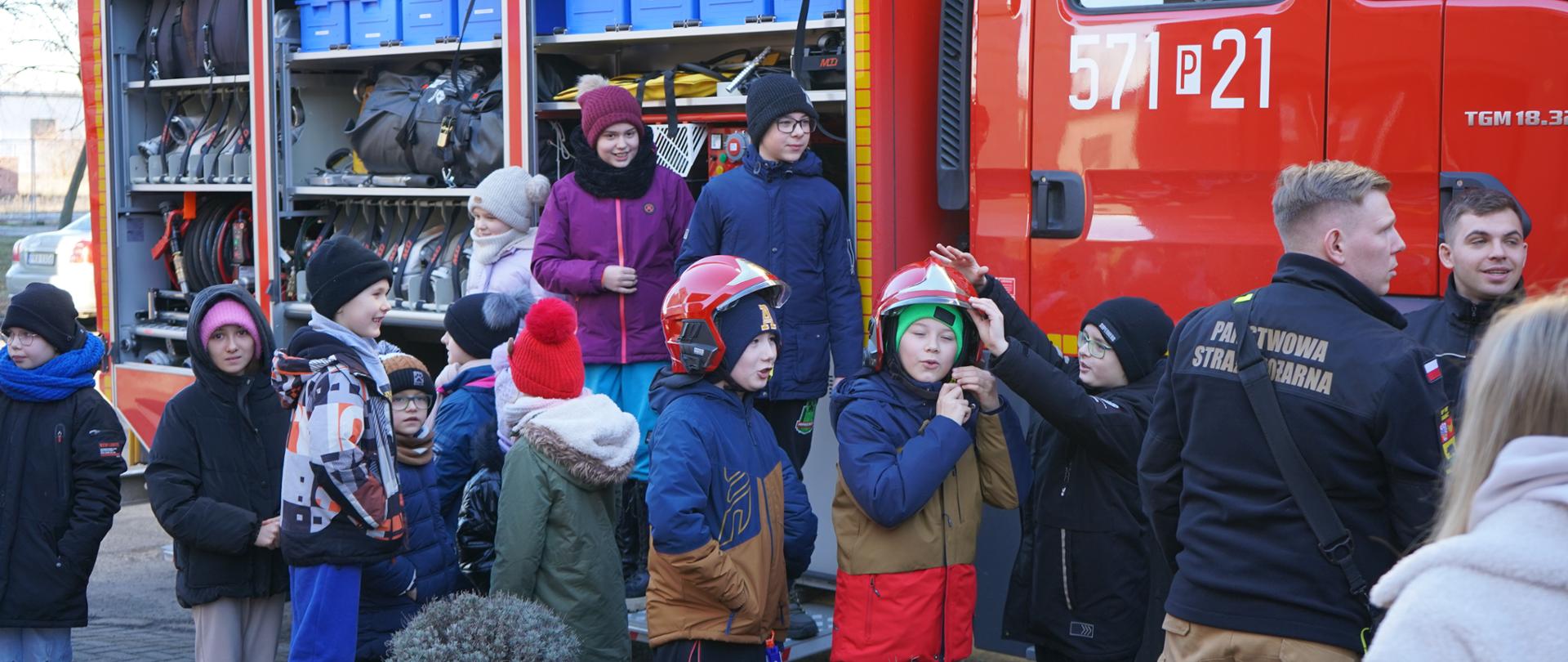 Plac przed Centrum Usług Społecznych w Rawiczu. Na ulicy zaparkowany jest wóz strazacki. Przy nim znajdują sie uczestnicy spotkania, którzy oglądają samochód i znajdujący sie na nim sprzęt. Dwóch chłopców ma załozone czerwone hełmy strażackie. 