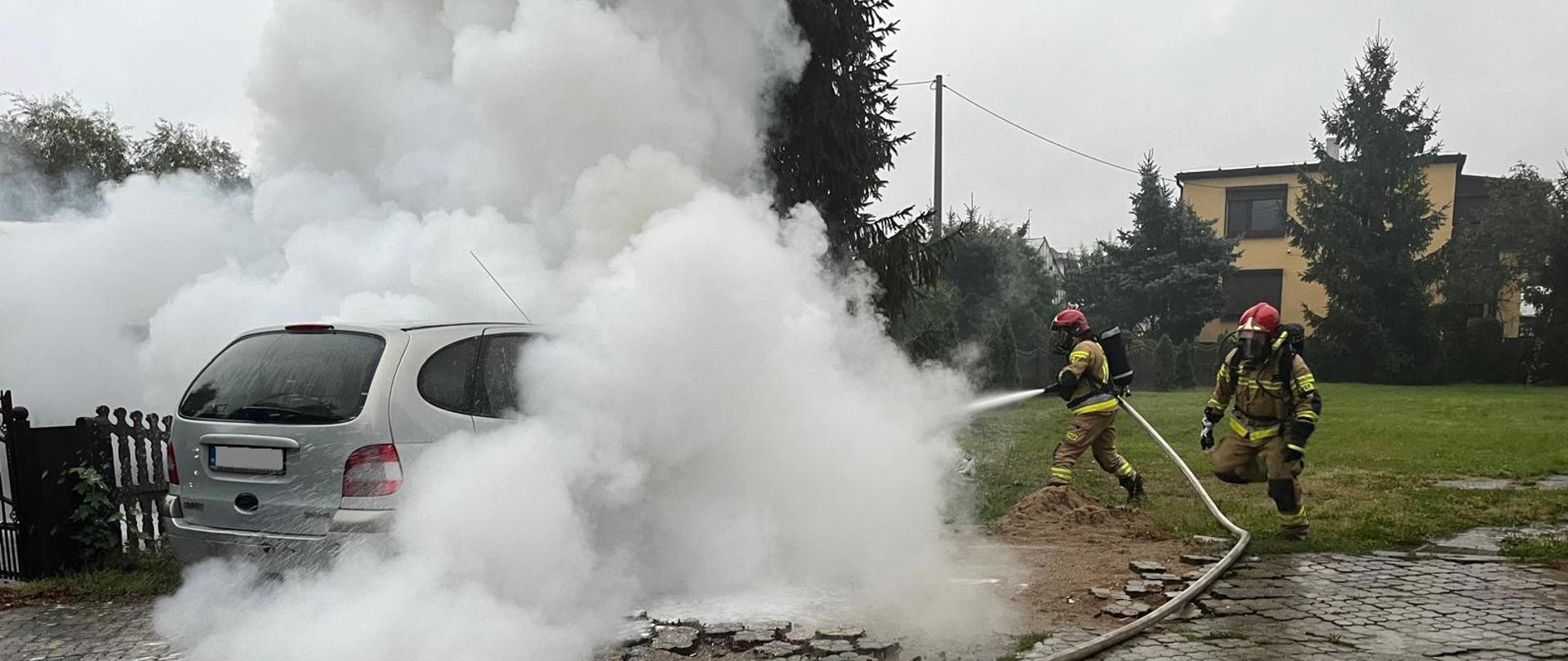 na posesji stoi w dużym zadymieniu samochód osobowy, przy nim strażak podaje prąd piany gaśniczej, obok niego biegnie kolejny strażak zabezpieczony aparatem dróg oddechowych