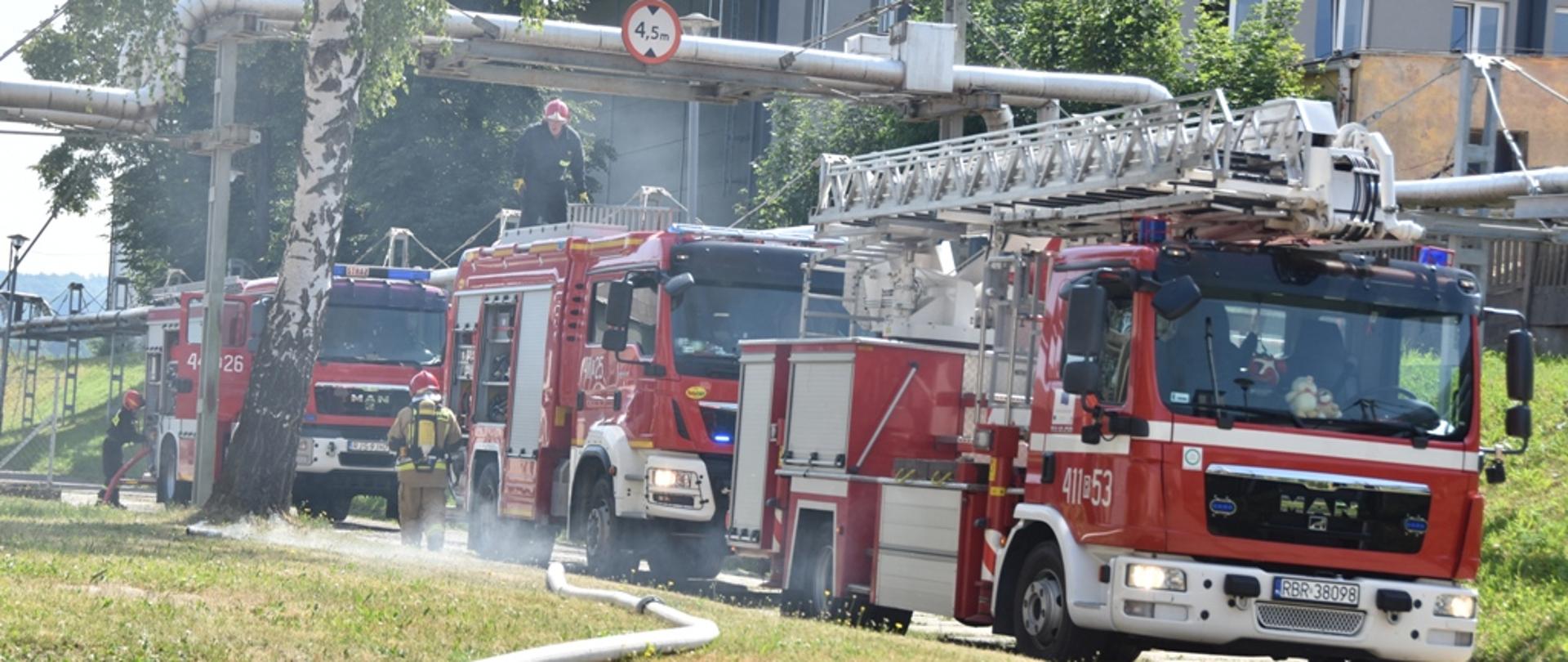 Kolorowa fotografia wykonana w słoneczny dzień na zewnątrz. Przedstawia ona ćwiczenia strażaków na terenie zakładu produkcyjnego.