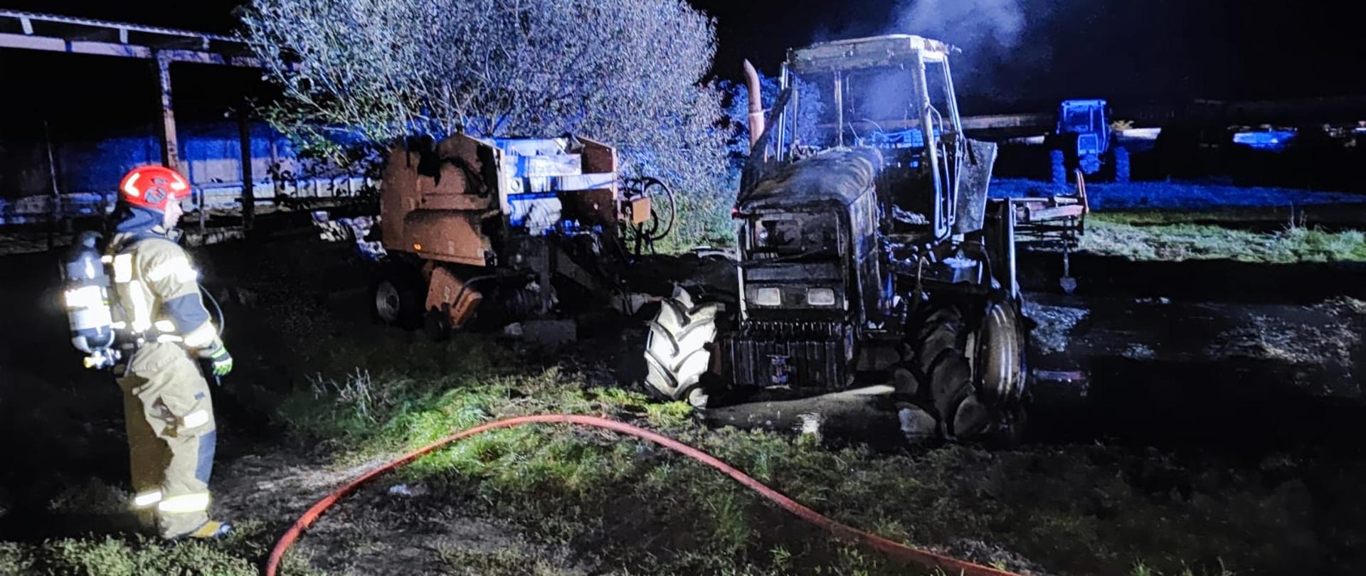 Pożar maszyn rolniczych, spalony ciągnik rolniczy i prasa do słomy, obok stoi strażak w czerwonym hełmie z założonym na plecach aparatem powietrznym, przy nim leży linia gaśnicza zakończona prądownicą.