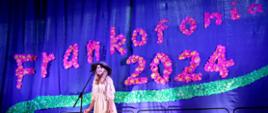 Dziewczynka ubrana w beżową sukienką i kapelusz, stoi przed mikrofonem. Z tyłu na fioletowym tle napis w kolorze różowym "Frankofonia 2024"".