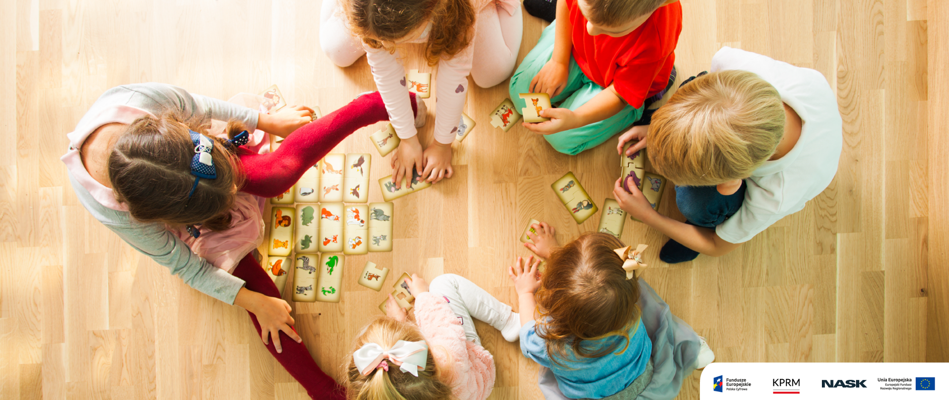 Zdjęcie przedstawia siedzące na podłodze dzieci, grające w grę. W prawym dolnym rogu logotypy: Fundusze Europejskie, KPRM, NASK, UE.