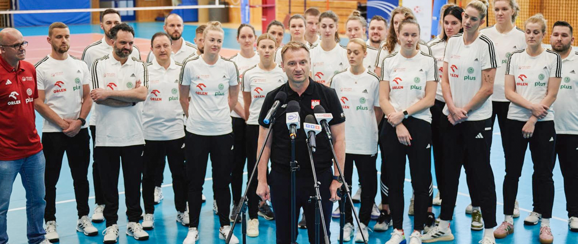 Konferencja Ministra Sportu i Turystyki w Szczyrku, 4 lipca 2024 r. Sławomir Nitras stoi przed mikrofonami w hali sportowej. Za nim stoją sportowcy. W tle widać m.in. siatkę do siatkówki.