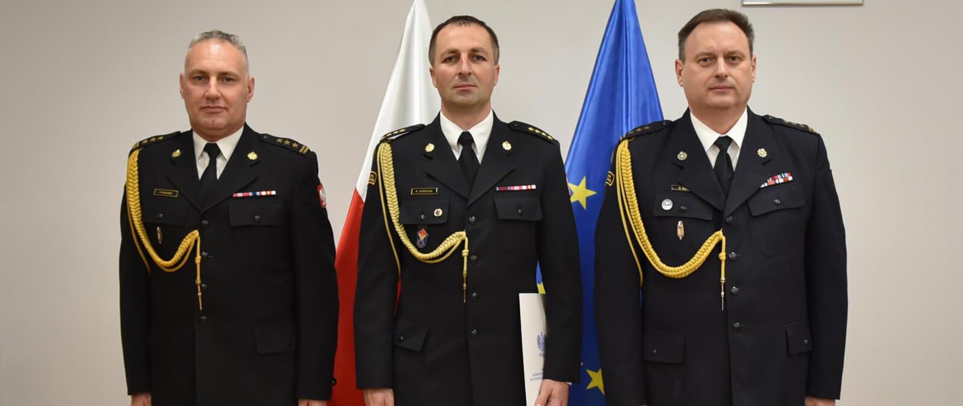 trzech strażaków pozujących do zdjęcia w gabinecie, za nimi flagi polski i unii europejskiej