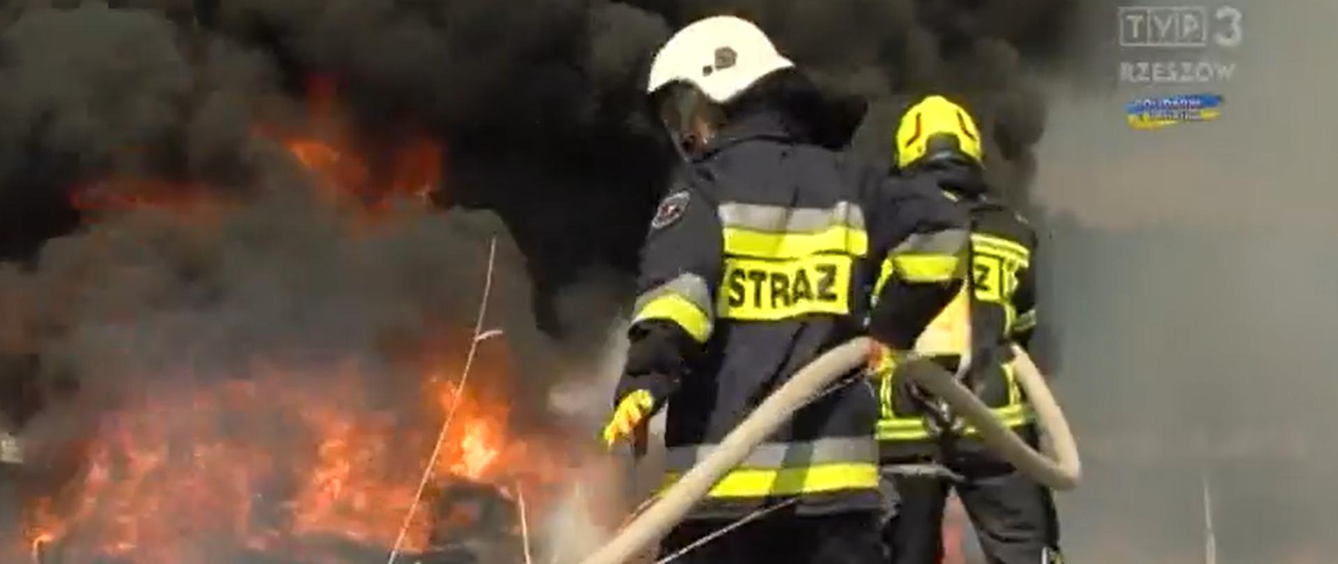 Zdjęcie przedstawia 2 strażaków w ubraniach specjalnych z linią wężową podczas gaszenia pożaru.