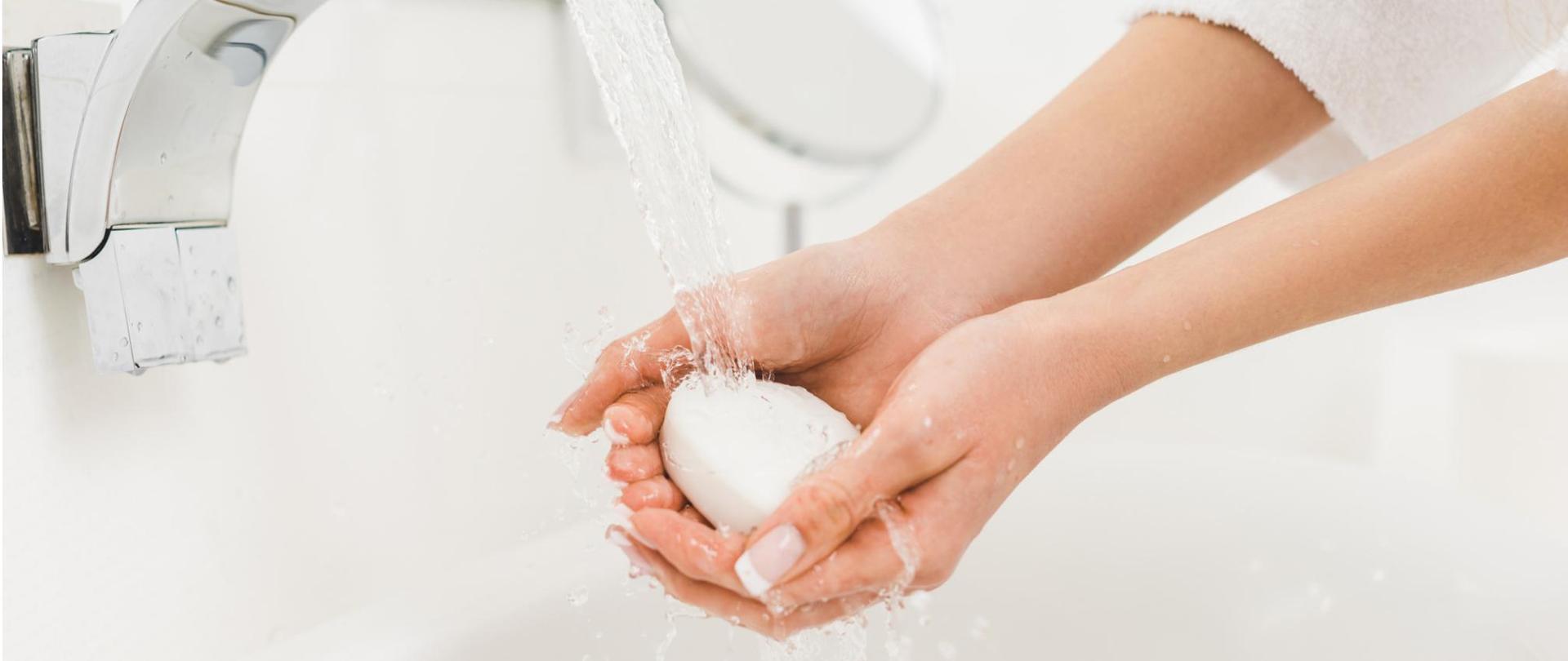 Na zdjęciu są widoczne dłonie kobiety, które trzyma pod strumieniem wody. W dłoniach ma mydło.