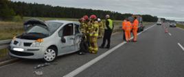 Droga ekspresowa S5. Na pasie awaryjnym stoi auto osobowe, które uczestniczyło w kolizji. Ma dość mocno uszkodzoną tylną część. Działania przy tym pojeździe prowadzą strażacy. Obok stoja pracownicy słuzby drogowej w pomarańczowych kombinezonach. Za nimi zaparkowana jest autolaweta. W oddali widać 2 pojazdy służby drogowej, które zabezpieczają teren oraz rozstawione stożki ostrzegawcze. Na jezdni odbywa się ruch kołowy. W tle łąka, lasy i wiadukt.