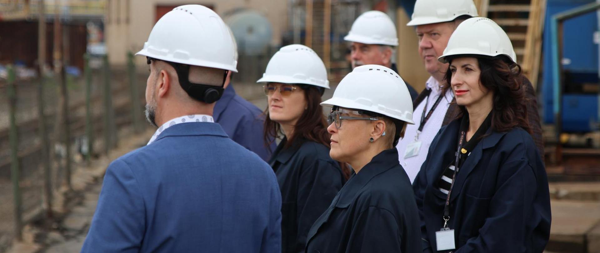 Wiceminister Mrówczyńska w białym kasku stoi wśród kilku osób w takich samych kaskach, patrzą w lewo, za nimi w tle widać wnętrze obiektu przemysłowego.