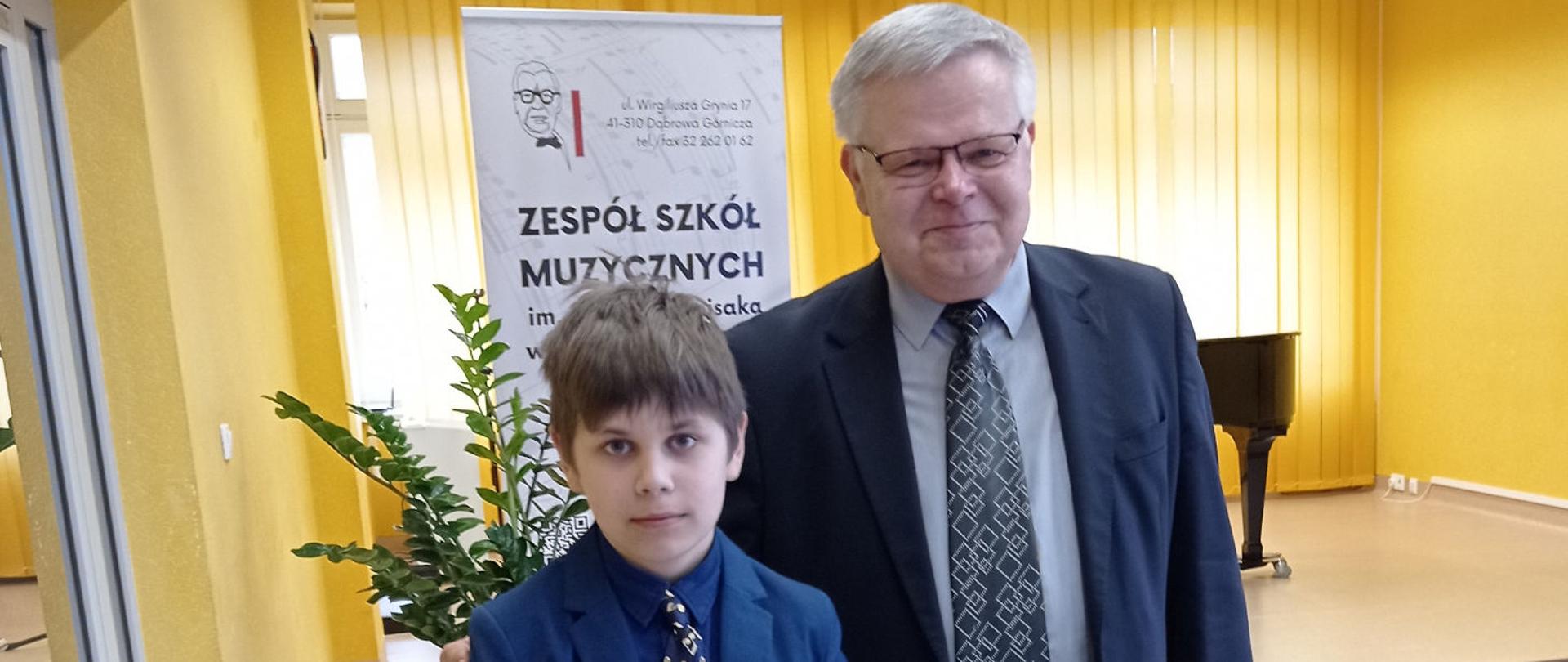 Na zdjęciu Julian Merecki z nauczycielem, w tle baner szkoły, żółte rolety.