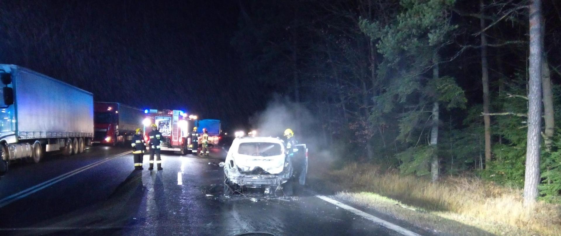 Zdjęcie przedstawia miejsce wypadku: z lewej strony stoją samochody ciężarowe, a po prawej spalony doszczętnie samochód osobowy, w głębi widać zaparkowany samochód gaśniczy i ratowników.