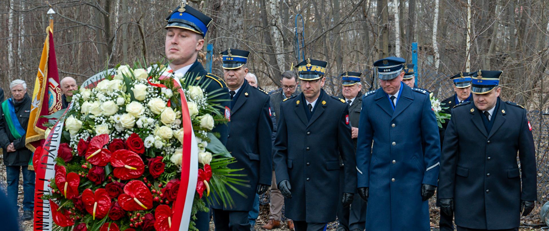 Przedstawiciele służb - policji i straży pożarnej. Na pierwszym planie funkcjonariusz z wieńcem z biało - czerwonych kwiatów