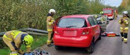 Zdjęcie przedstawia dwa samochody osobowe po zderzeniu stojące na drodze. Strażacy udzielają kwalifikowanej pierwszej pomocy poszkodowanym i przygotowują sprzęt hydrauliczny do wykonania dostępu do poszkodowanego kierowcy. W tle samochody pożarnicze i drzewa.