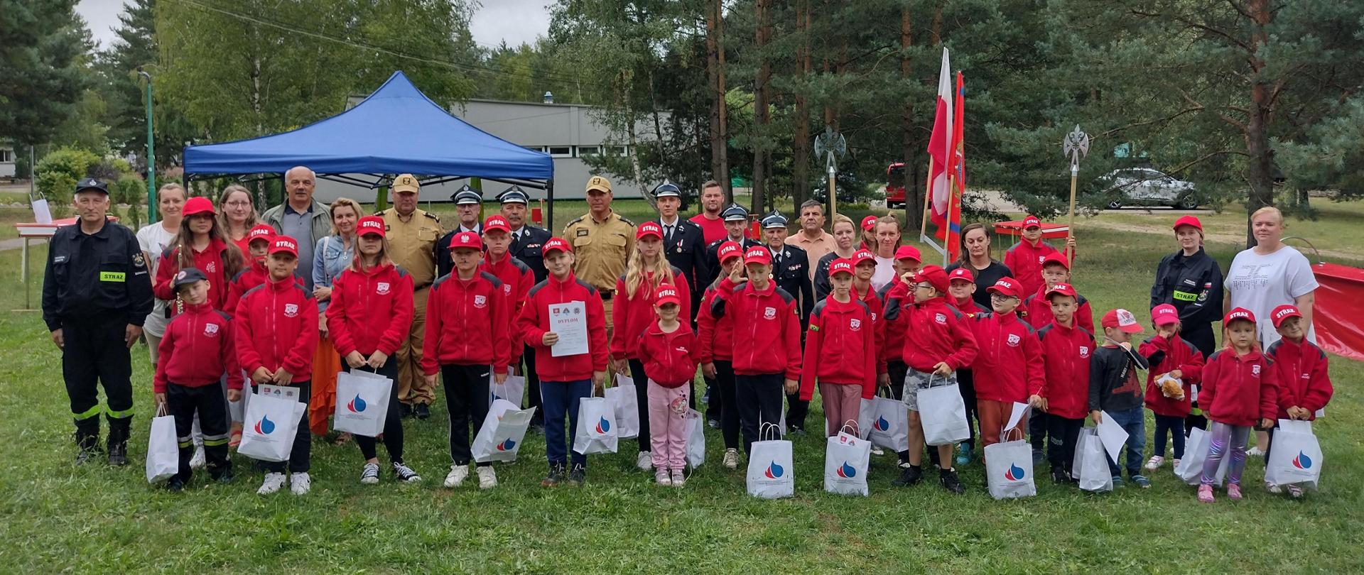 zdjęcie grupowe dzieci strażaków i opiekunów Wszyscy stoją na trawie dzieci trzymają białe torby z upominkami