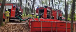 Dwa samochody pożarnicze stoją w lesie przy czerwonym zbiorniku na wodę. Kilku strażaków robi coś przy samochodach. Widoczne suche liście na ziemi i Wysokie drzewa w oddali. 