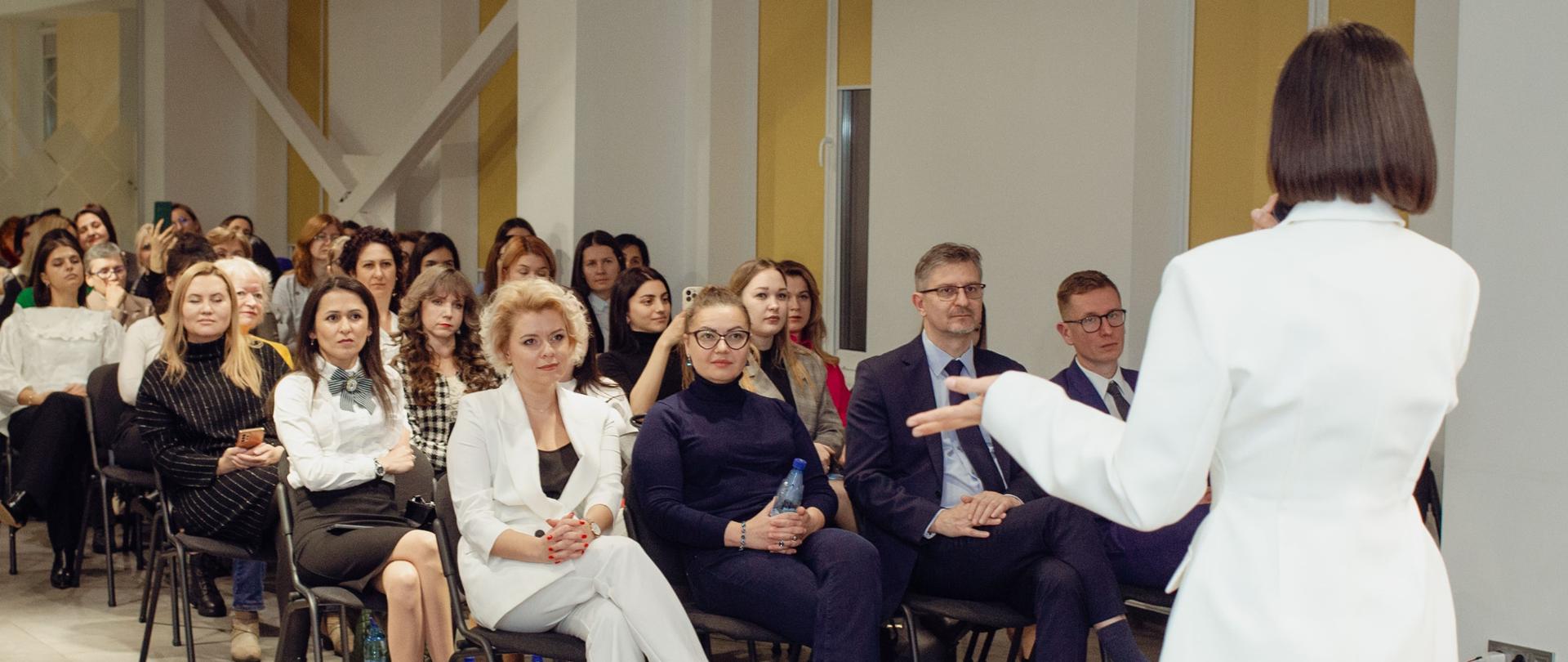 Sala konferencyjna. Kobiety siedzą na krzesłach ustawionych w rzędach. Jedna kobieta stoi przed nimi i przemawia do mikrofonu. 