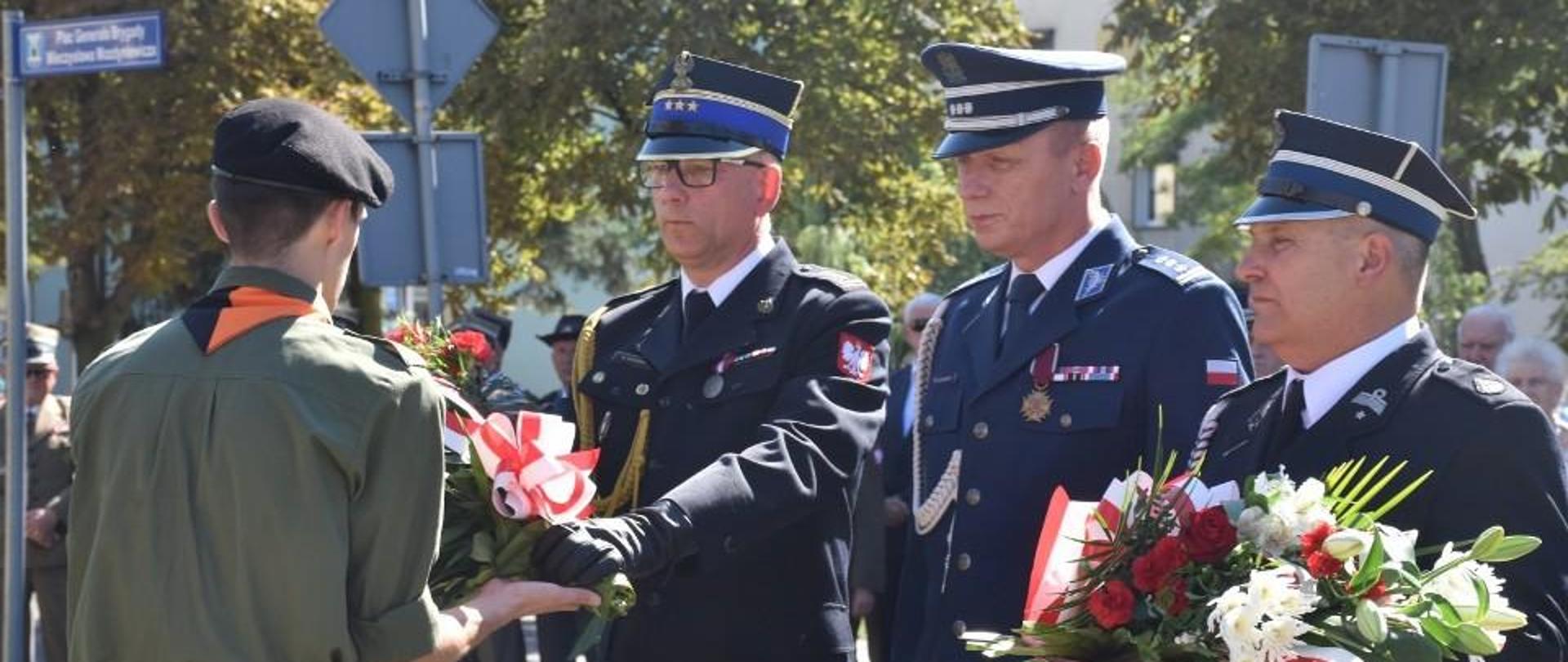 Trzech mężczyzn ubranych w mundury wyjściowe Państwowej Straży Pożarnej, Policji i Ochotniczej Straży Pożarnej stoi obok siebie, frontem do nich stoi harcerz, któy odbiera wieniec kwiatów od mężczyzny w mundurze Państowej Straży Pożarnej