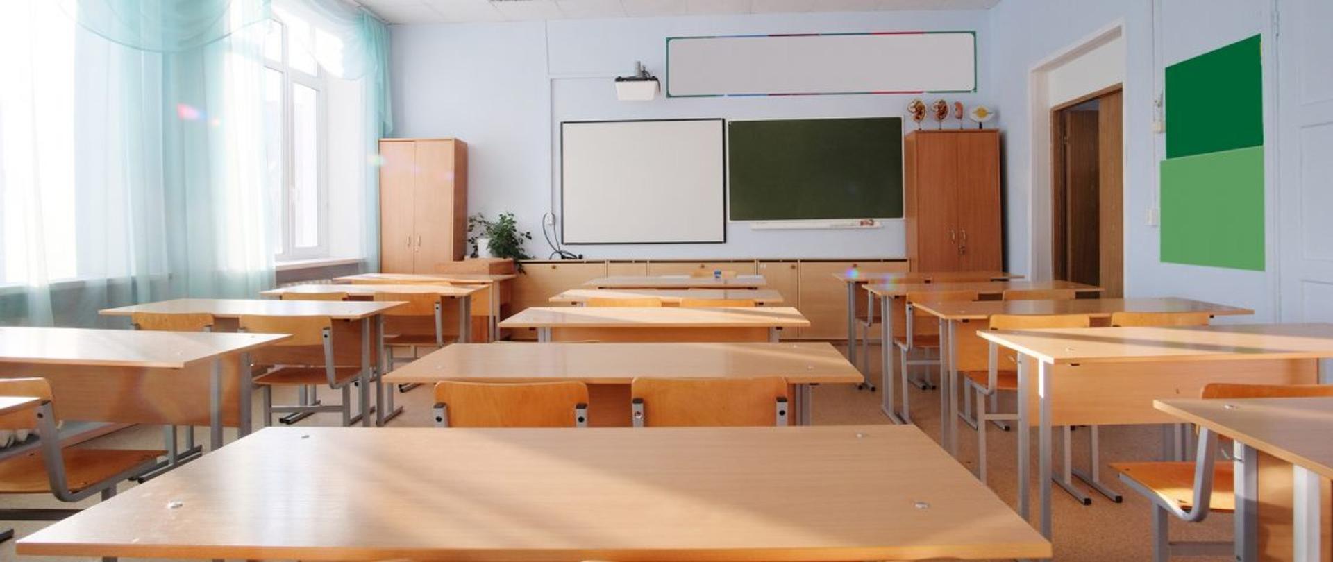 sala lekcyjna w szkole z ustawionymi w trzech rzędach ławkami na wprost których na ścianie wiszą dwie tablice 