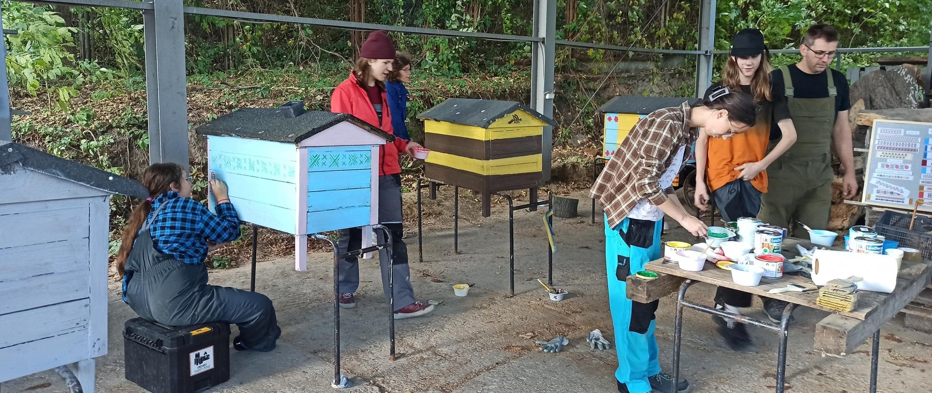 Uczniowie malują ule. Na stole leżą przybory malarskie, np. puszki z farbą, pędzle, rolka papieru do wycierania, pojemniki, itd. W tle pniaki drzew i roślinność.