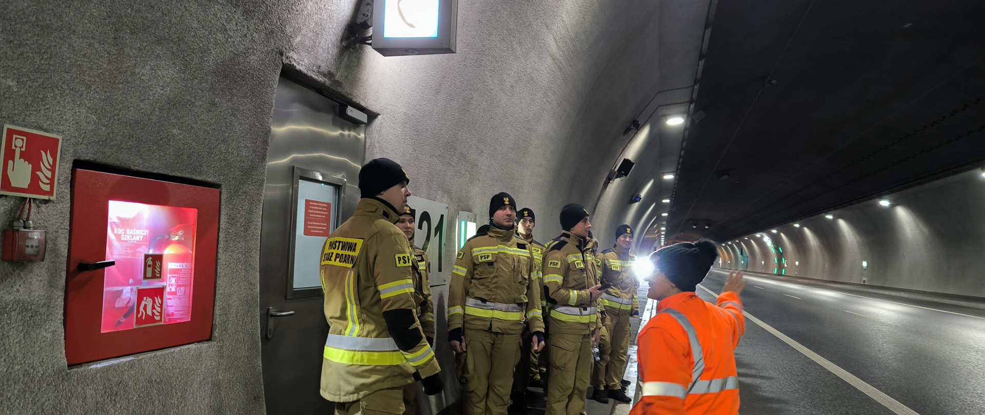 Wnętrze tunelu drogowego wraz z widokiem jego infrastruktury. Wewnątrz grupa ubranych w mundury koloru piaskowego strażacy. 