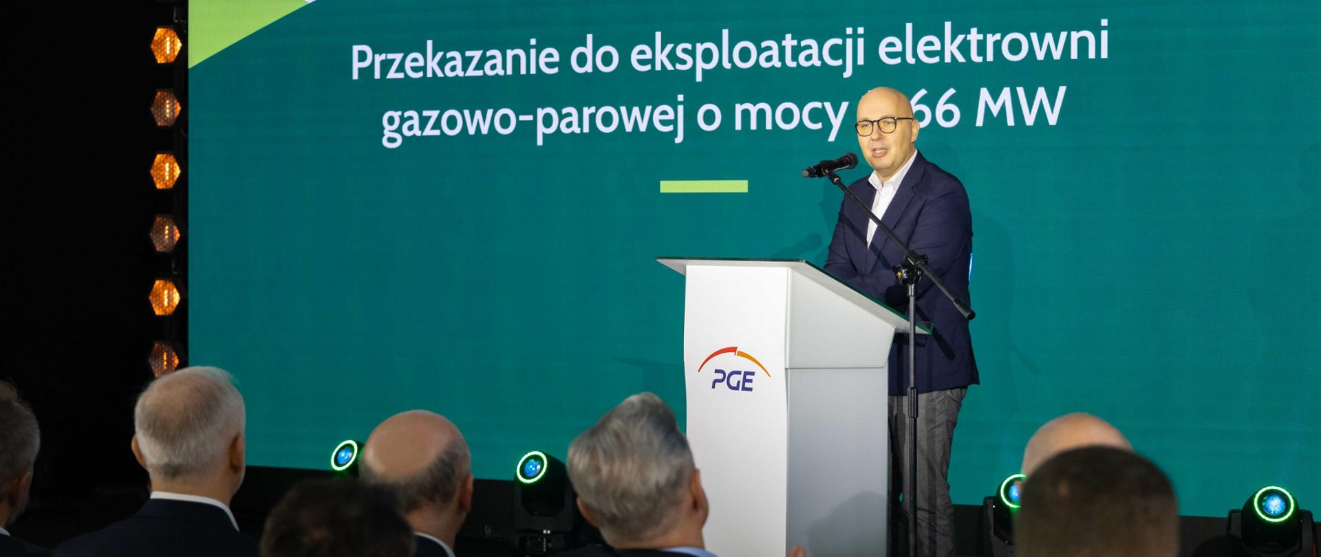 Wiceminister Robert Kropiwnicki stoi za mównicą. W tle ekran z napisem Przekazanie do eksploatacji elektrowni gazowo-parowej o mocy 1366 MW. Ujęcie zza słuchaczy.