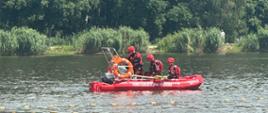 Czterech strażaków w łodzi ratowniczej w czerwonych skafandrach płynie po wodzie. Za nimi w tle zarośla i drzewa liściaste. Przed nimi lina pływająca wyznaczająca kąpielisko.