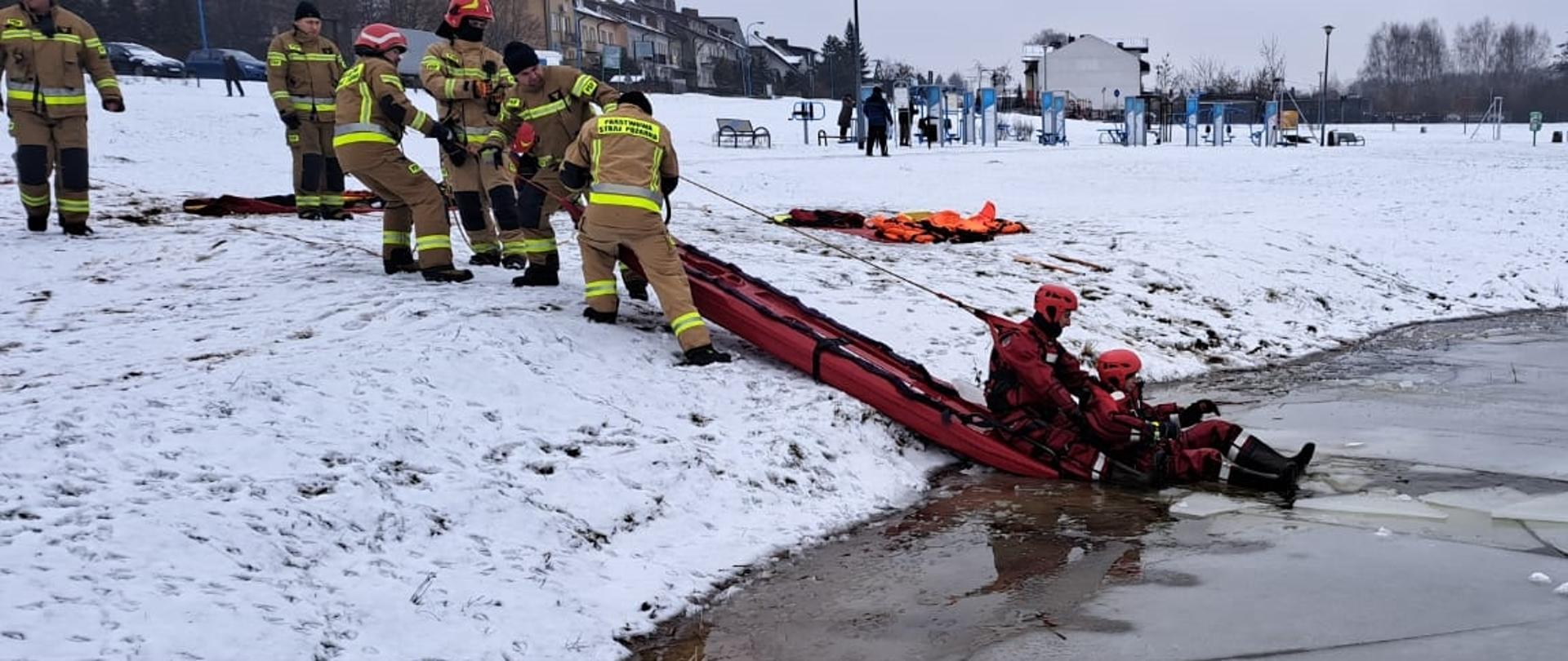 Brzeg wody. Strażak-ratownik na saniach lodowych z poszkodowanym, pozostali ratownicy wyciągają sanie na brzeg.