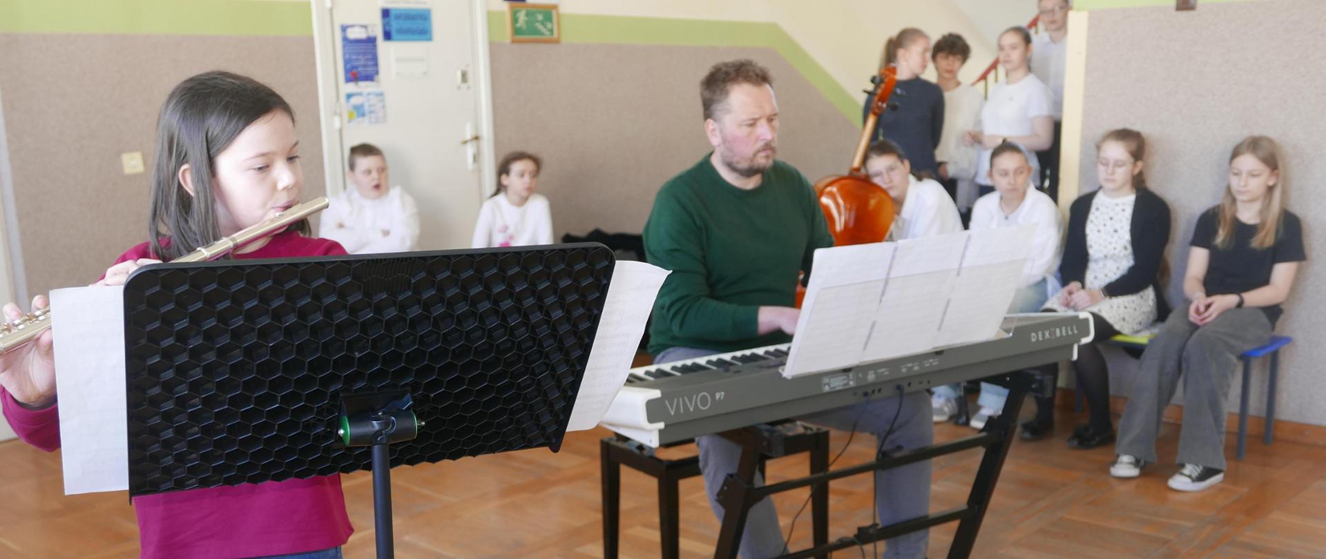 Na pierwszym planie dwójka instrumentalistów. Dziewczynka grająca na flecie poprzecznym lekko schowana za pulpitem nutowym, a obok pan akompaniujący jej na pianinie cyfrowym. Za nimi czekająca na swoją kolej by wystąpić młodzież .