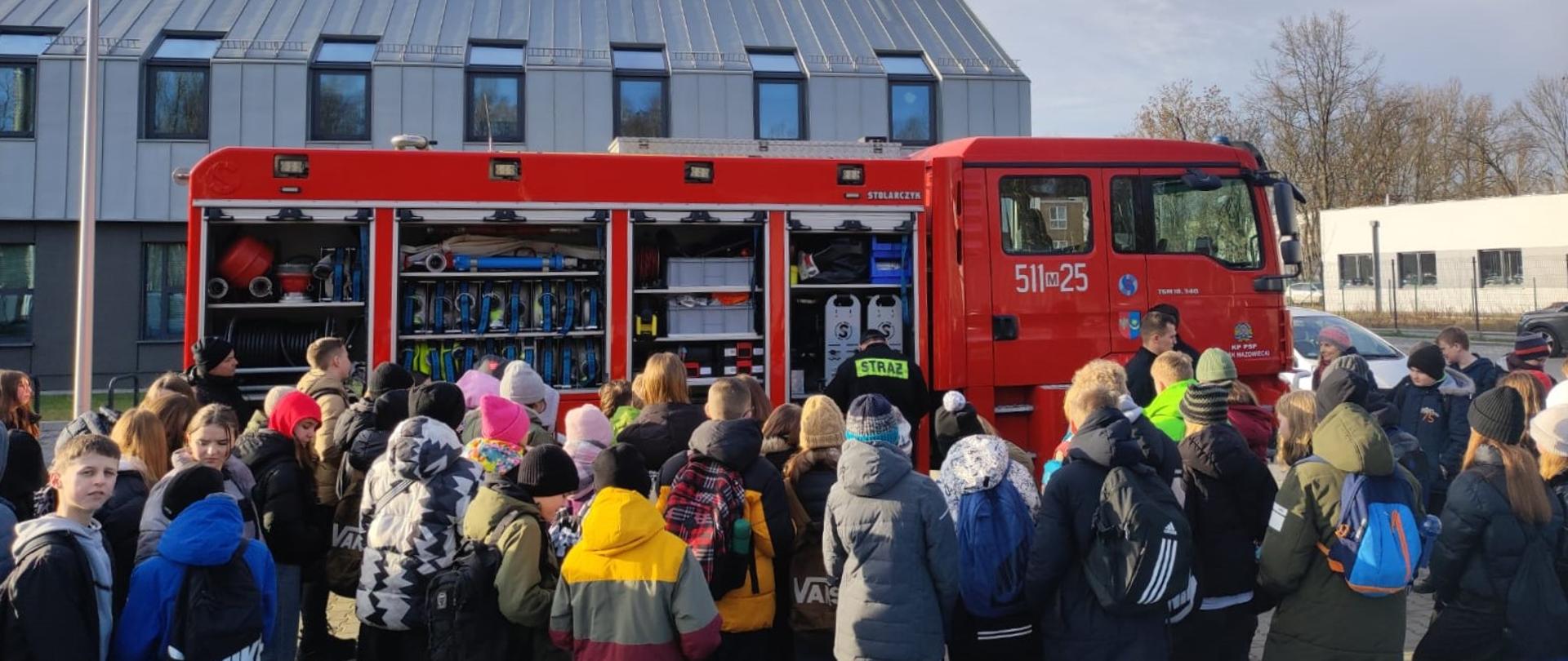 Spotkanie z dziećmi i młodzieżą w ramach kampanii edukacyjnej "Bezpieczne ferie" pod budynkiem KPP w Mińsku Mazowieckim. Zebrana młodzież stoi wokół samochodu ratowniczo-gaśniczego i ogląda sprzęt, który znajduje się w skrytkach samochodu oraz ten który prezentują strażacy.