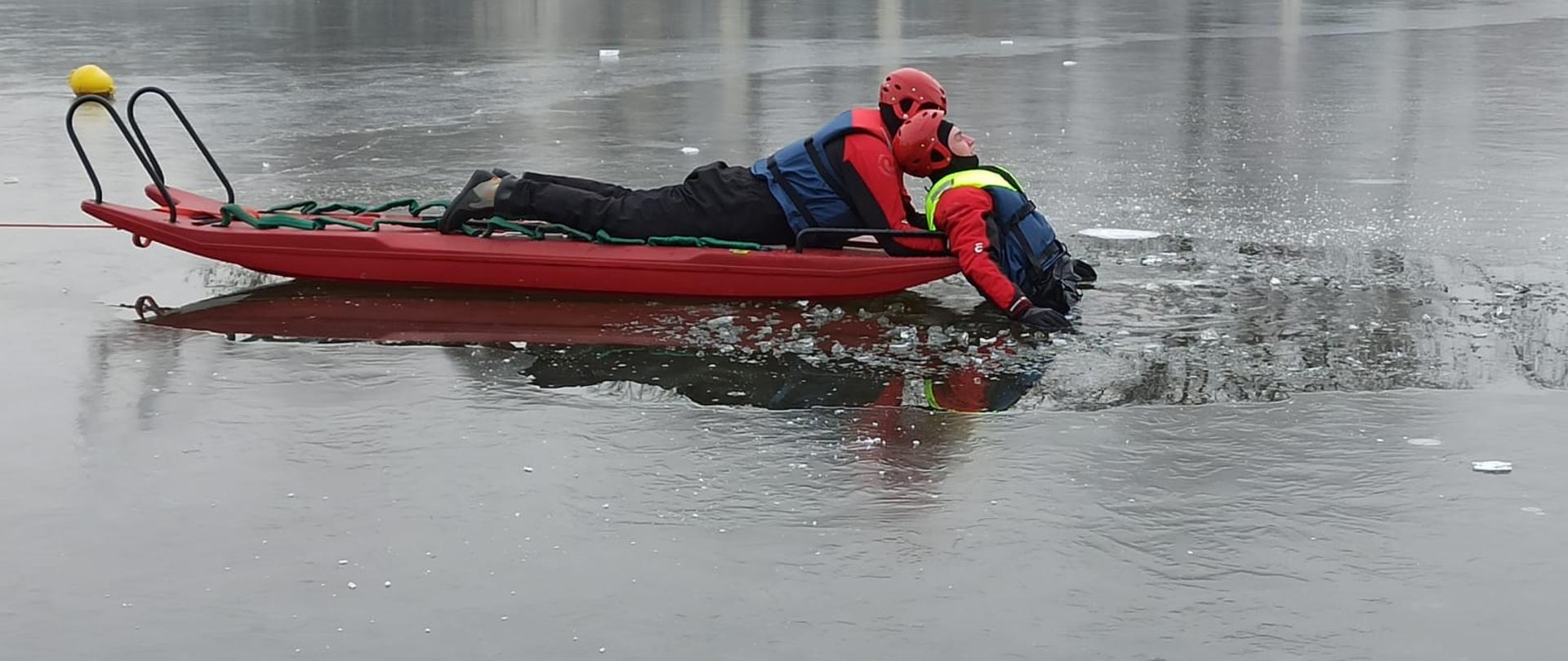 Zdjęcie przedstawia dwóch strażaków w ubraniach wypornościowych, którzy przy użyciu sani lodowych ćwiczą podejmowanie osoby poszkodowanej (topielca) z akwenu.