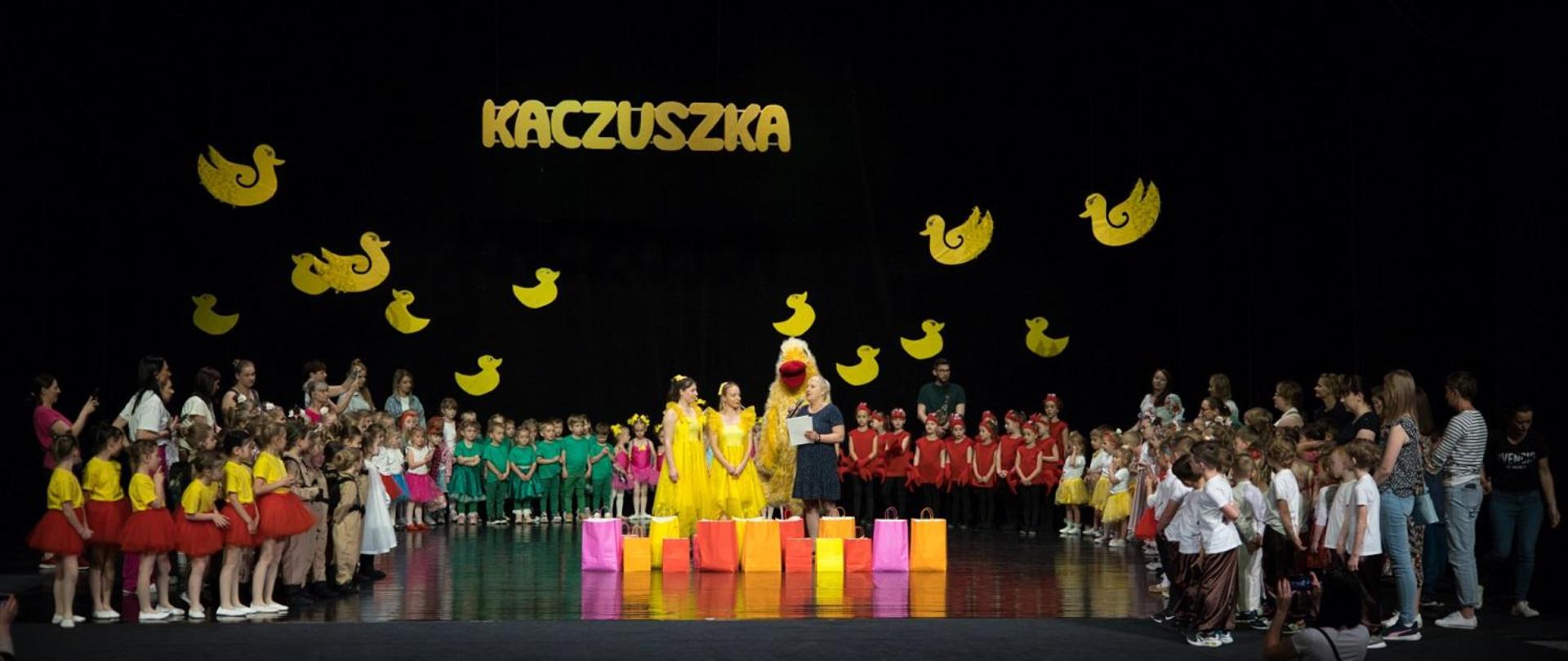 Zdjęcie przedstawia stojących na scenie uczestników Festiwalu Tańczących Dzieci „Kaczuszka”. Na górze sceny jest żółty napis Kaczuszka, obok są żółte kaczki. 