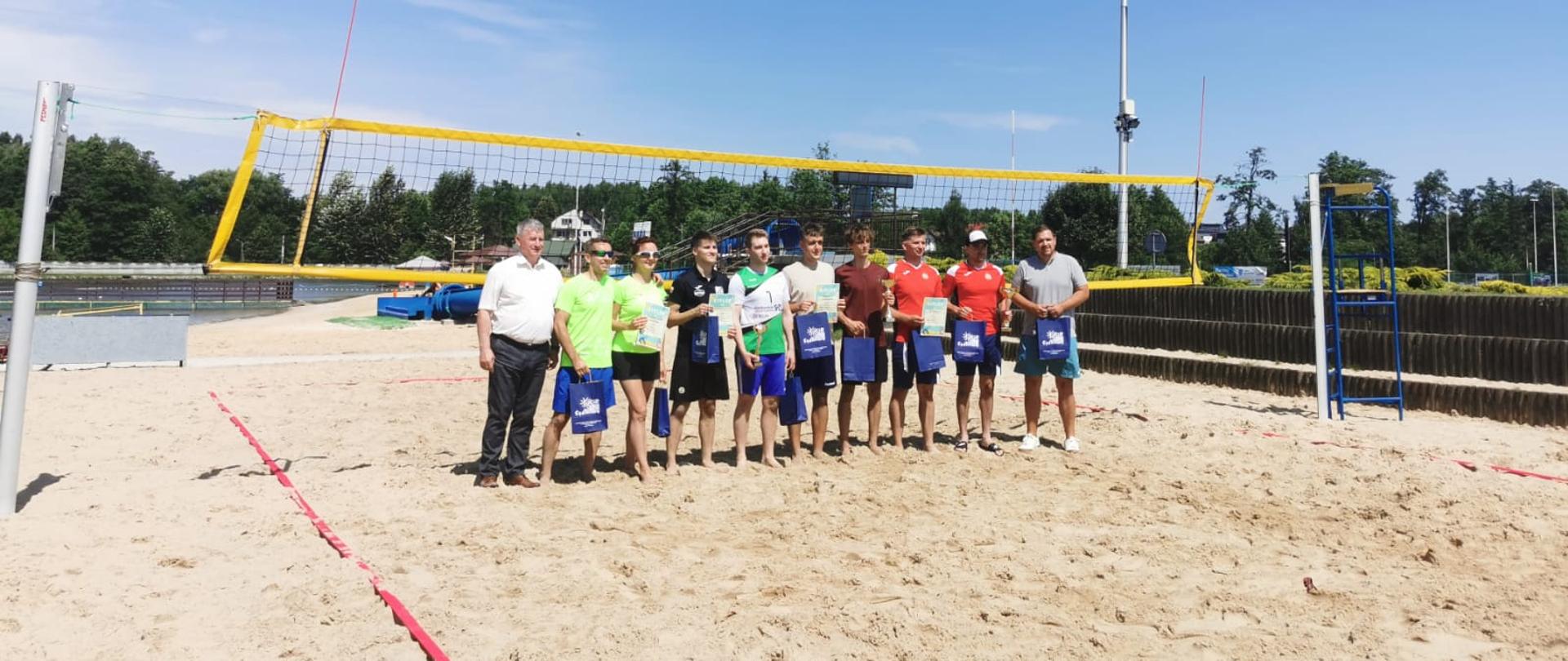 Zdjęcie przedstawia wszystkie drużyny podczas XVI Gminnego Otwartego Turnieju Siatkówki Plażowej. Zdjęcie zrobione na tle siatki na boisku do siatkówki plażowej. Zawodnicy trzymają puchary oraz nagrody rzeczowe.