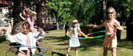 Zdjęcie kolorowe. Cztery dziewczynki w letnich strojach uprawiają gimnastykę na trawniku. W tle zielone drzewa.