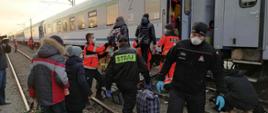 Na torach stoi pociąg, przy nim ratownicy medyczni i strażacy, którzy pomagają wsiadać do pociągu kilku osobom cywilnym.