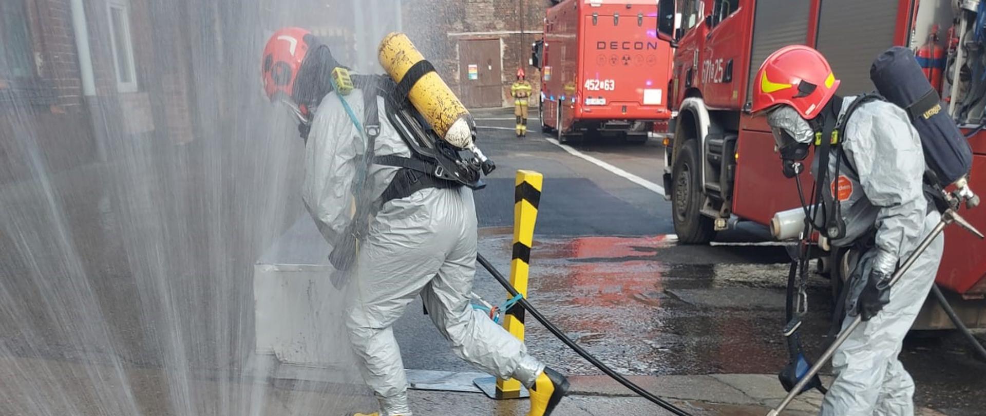 Strażacy w kombinezonach chemoodpornych przechodzą przez kurtynę wodną do strefy, gdzie doszło do wycieku amoniaku.