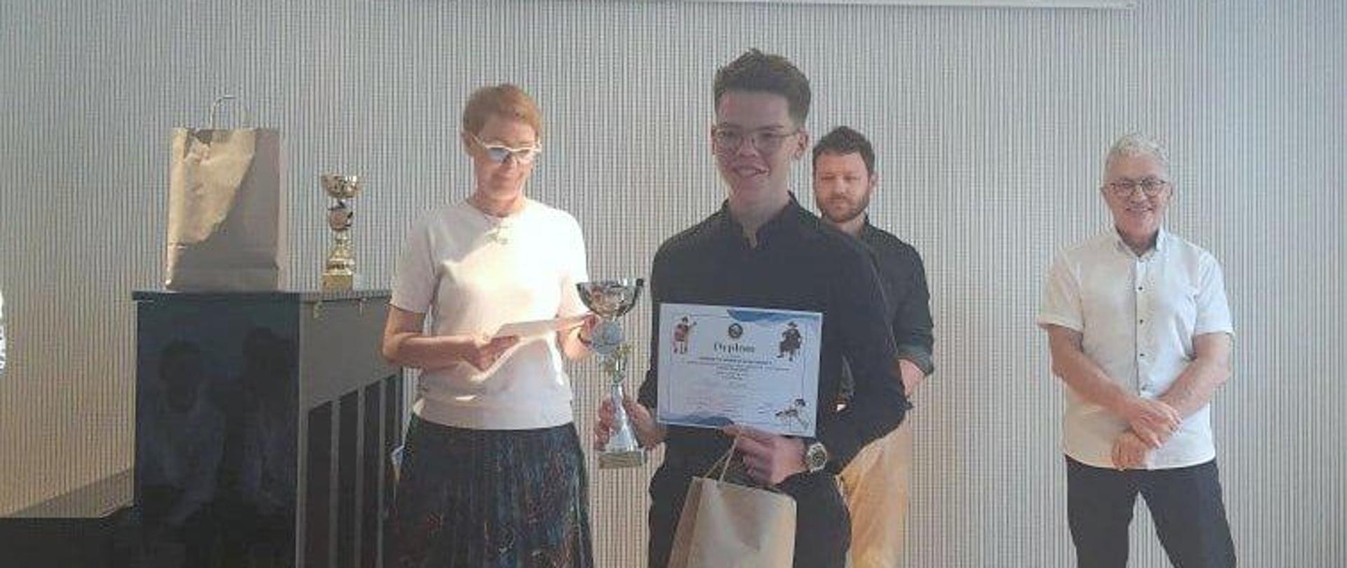 Uczeń trzymający puchar i dyplom, za nim trzy osoby (jury konkursu).