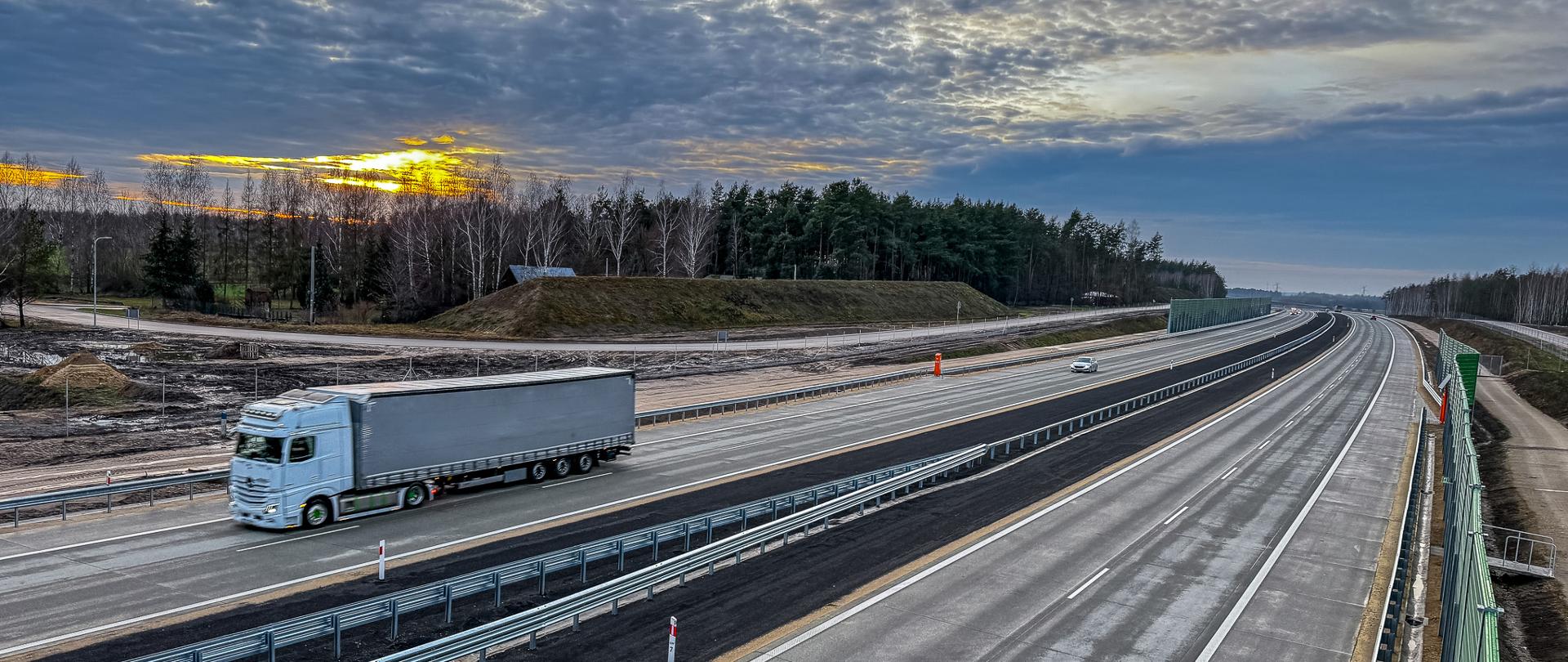 Zdjęcie przedstawia szeroką autostradę z kilkoma pasami ruchu w obu kierunkach. Na lewym pasie widoczna jest ciężarówka, a na prawym pasie, w oddali, znajduje się samochód osobowy. Tło stanowi las oraz zachodzące słońce, które tworzy piękne, złote światło na niebie pokrytym chmurami. Po obu stronach autostrady znajdują się barierki ochronne oraz ekrany dźwiękochłonne.