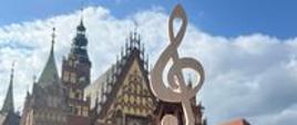 Zdjęcie przedstawia budynki miasta Wrocław na drugim planie, na pierwszym planie kilka osób oraz ręka z symbolem klucza wiolinowego