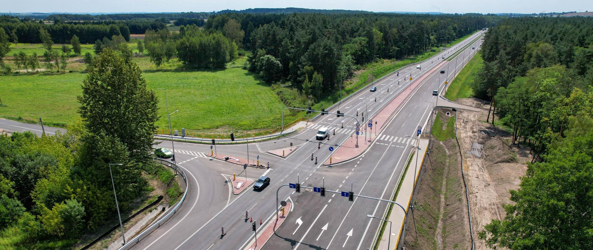 Na zdjęciu widzimy lotniczy widok na odcinek przebudowanej drogi krajowej nr 91 Markowice-Brudzowice oraz mniejszą drogę, która krzyżuje się z nią. Droga ma dwie jezdnie i wiele pasów ruchu w obu kierunkach, oddzielonych środkowym pasem z barierami. Na drodze poruszają się pojazdy, w tym samochody i ciężarówki, co wskazuje na ruch drogowy. Po obu stronach drogi znajduje się bujna zieleń z drzewami i trawiastymi obszarami. Niebo jest bezchmurne z niewielką ilością chmur, sugerując dobre warunki pogodowe.
To interesujące zdjęcie ukazuje infrastrukturę drogową i jej integrację z otaczającą przyrodą i zabudową.