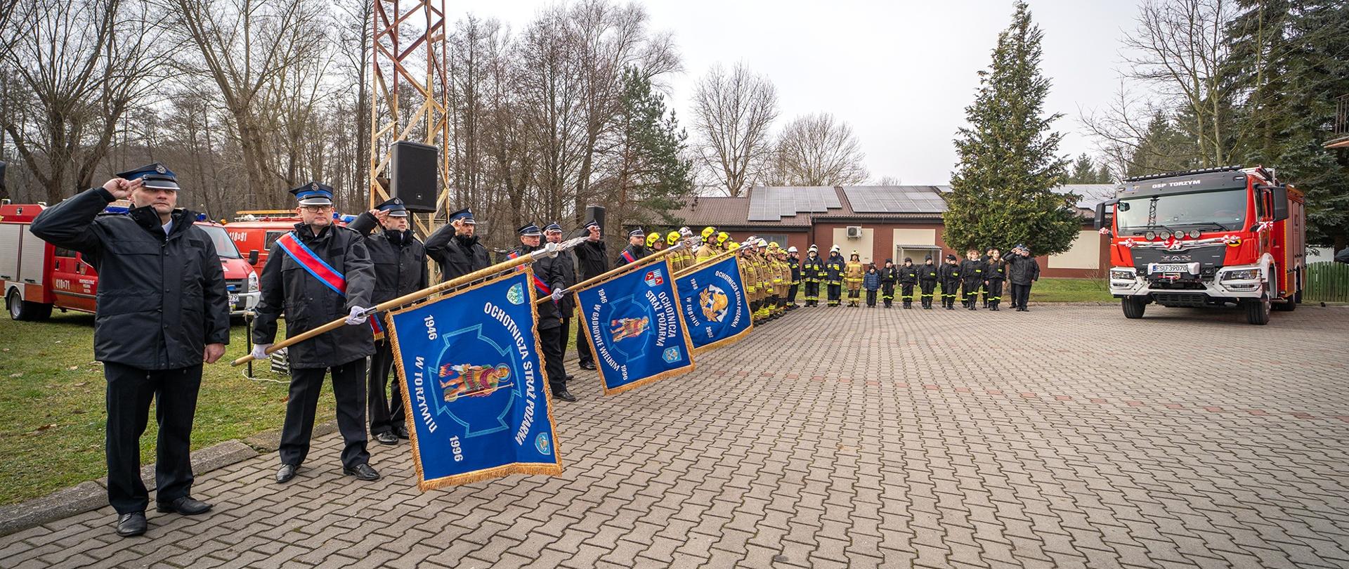 Poczty sztandarowe w szyku oddające honor, po prawej samochód pożarniczy