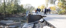 Zapadnięta i zniszczona droga w jednej z miejscowości w powiecie głubczyckim. W tle widać uczestników wizji terenowej, który idą obok zapadniętych fragmentów drogi. 