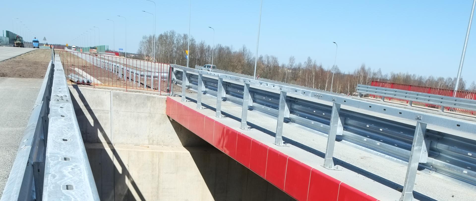 Fotografia przedstawia dwa równoległe obiekty inżynierskie. Wyposażone są w mostowe bariery ochronne. Cokół pomostu został wykonany w kolorze czerwonym.
