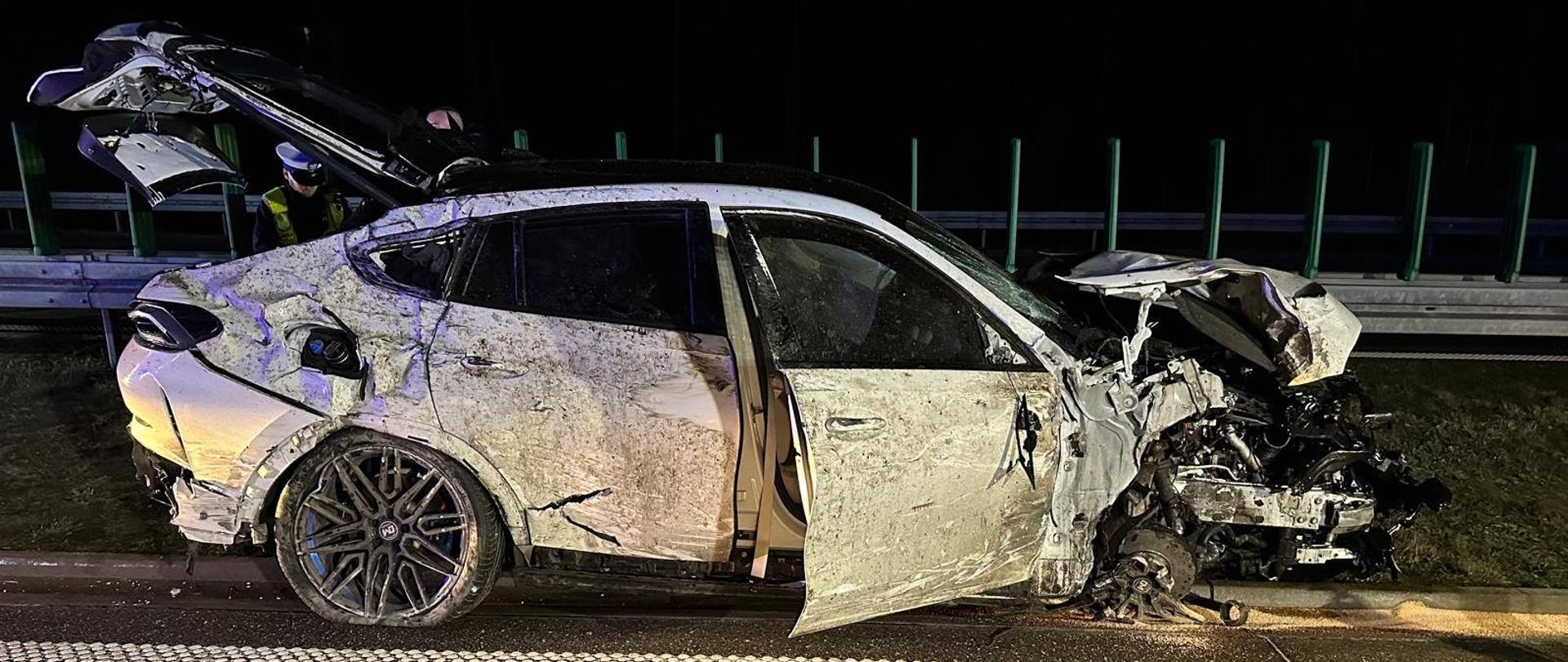 Samochód osobowy po dachowaniu, widok z boku, zniszczona karoseria, otwarte drzwi pasażera i klapa bagażnika. 