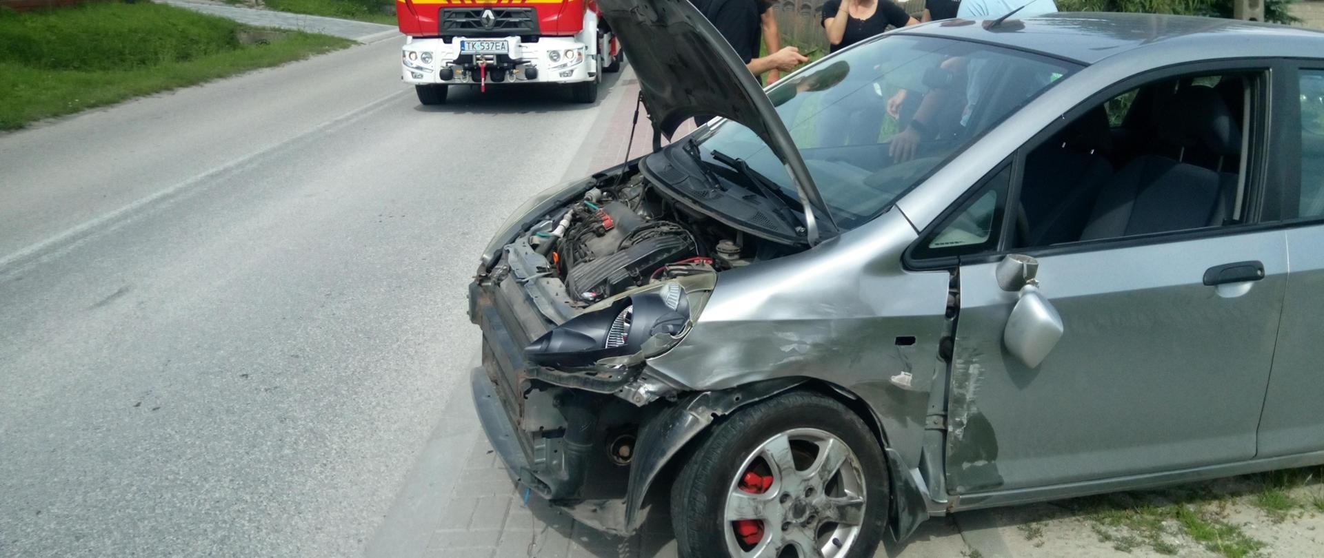 Zdjęcie przedstawia uszkodzony samochód po wypadku stojący na poboczu. Za nim po lewej stronie widać stojący samochód pożarniczy.