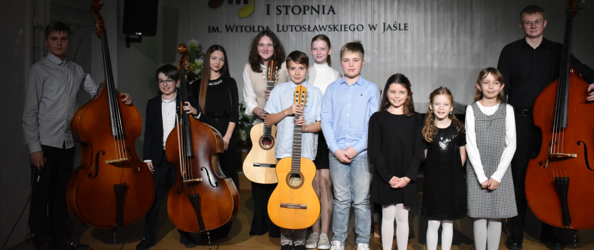 Na zdjęciu znajduje się grupa uczniów, niektórzy z instrumentami - gitarami i kontrabasami, w tle na jasnej ścianie podświetlonej zielonym kolorem, logo szkoły i godło państwowe 