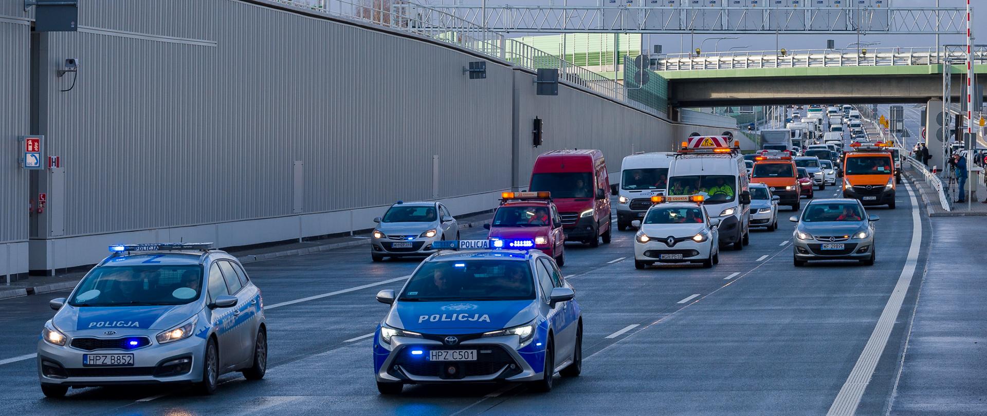 Zdjęcie przedstawia scenę na wielopasmowej drodze z wieloma pojazdami, w tym dwoma radiowozami z migającymi niebieskimi światłami, czerwonym vanem oraz kilkoma innymi samochodami i pojazdami serwisowymi GDDKiA. Wszystkie pojazdy poruszają się za dwoma radiowozami na pierwszym planie.