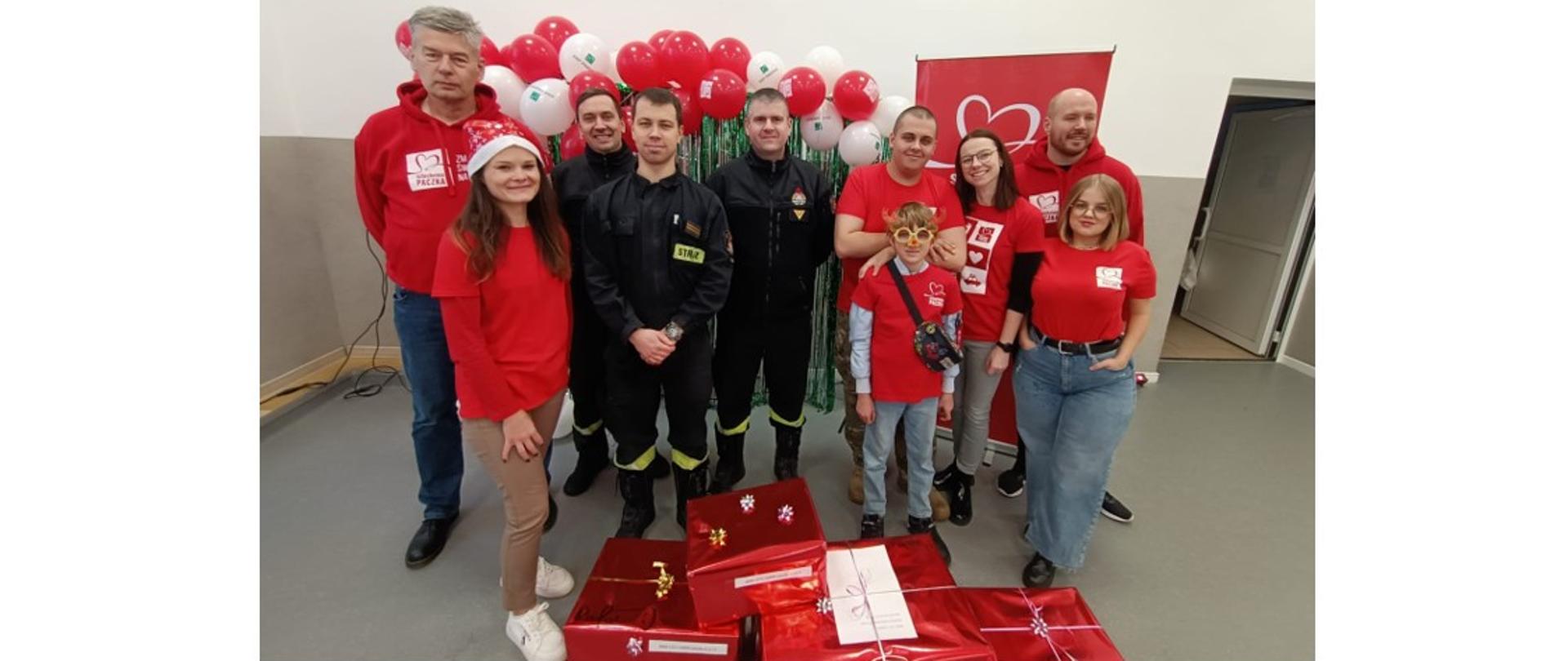 Zdjęcie przedstawia trzech funkcjonariuszy Komendy Powiatowej Państwowej Straży Pożarnej w Gołdapi oraz siedmiu Wolontariuszy akcji "Szlachetna Paczka" stojących obok przekazywanych w ramach przedsięwzięcia przez strażaków upominków, w siedzibie kwatery głównej akcji „Szlachetna Paczka” w powiecie gołdapskim